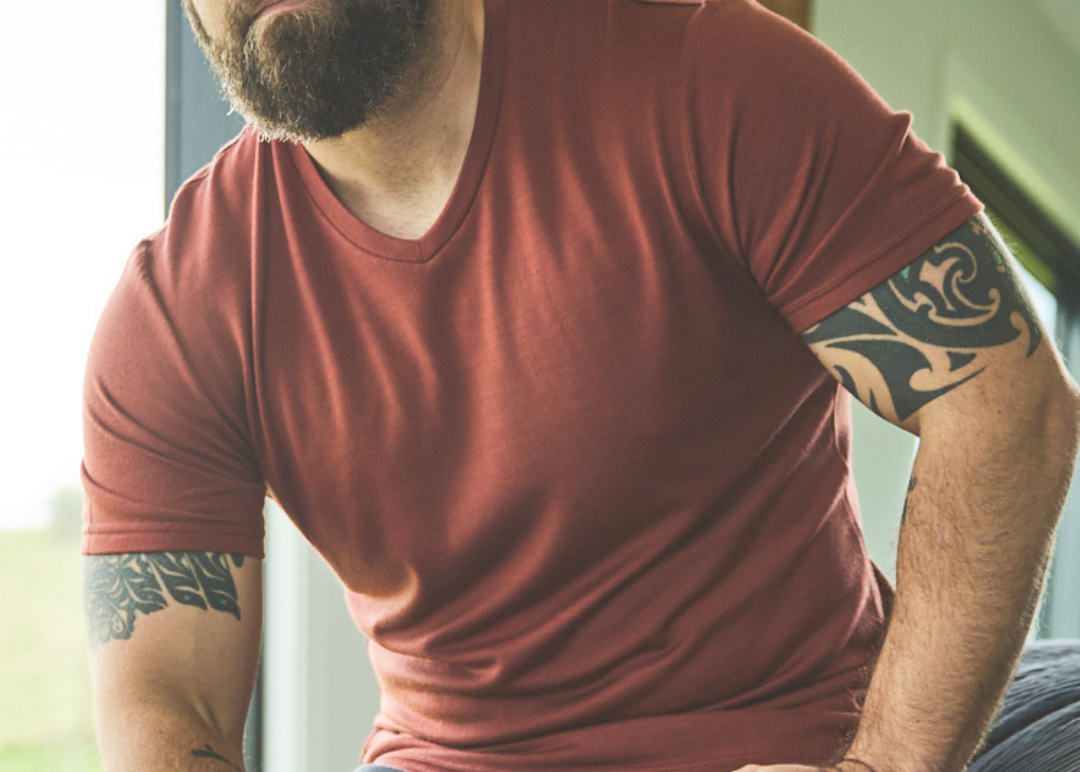 Man with tattoos wearing a red merino top sitting indoors.